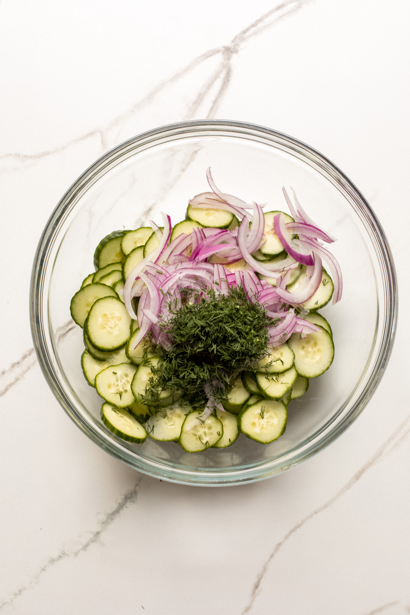 sliced cucumbers, onions, and dill in a glass mixing bowl.